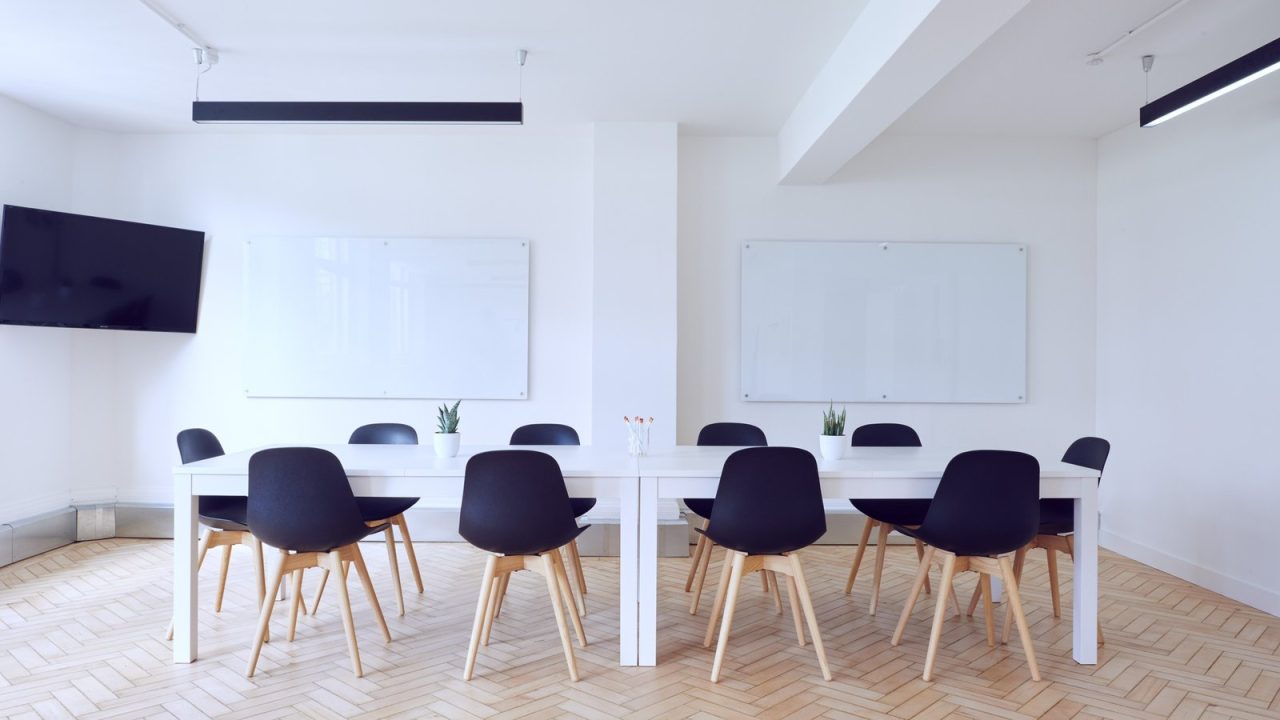 Salle de réunion lumineuse avec tables blanches, chaises noires, téléviseur et tableaux muraux