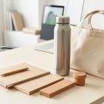 Assortiment élégant d’objets publicitaires durables disposés sur un bureau professionnel lumineux, comprenant une gourde en acier, un carnet vierge, un sac en coton et des accessoires en liège.